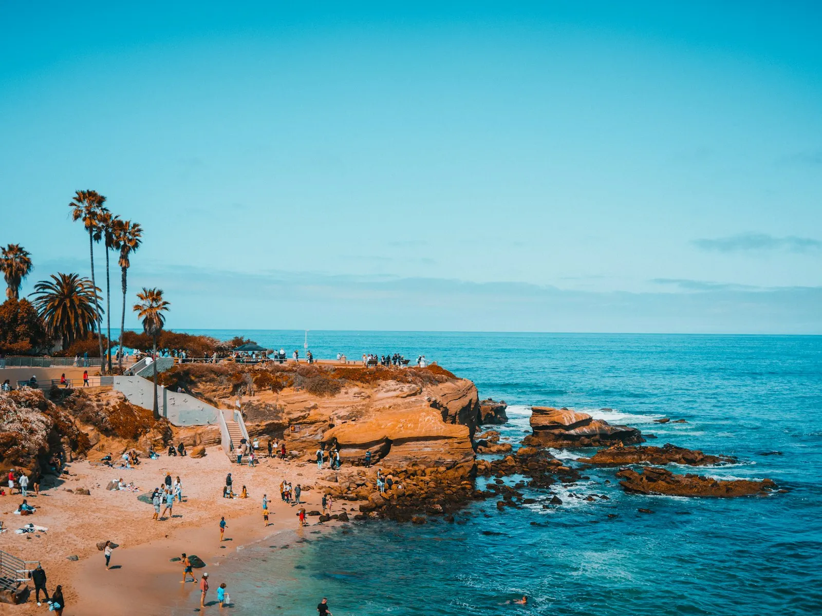 Enseada de La Jolla com mar azul-turquesa, rochas douradas, palmeiras e banhistas pequenos na areia