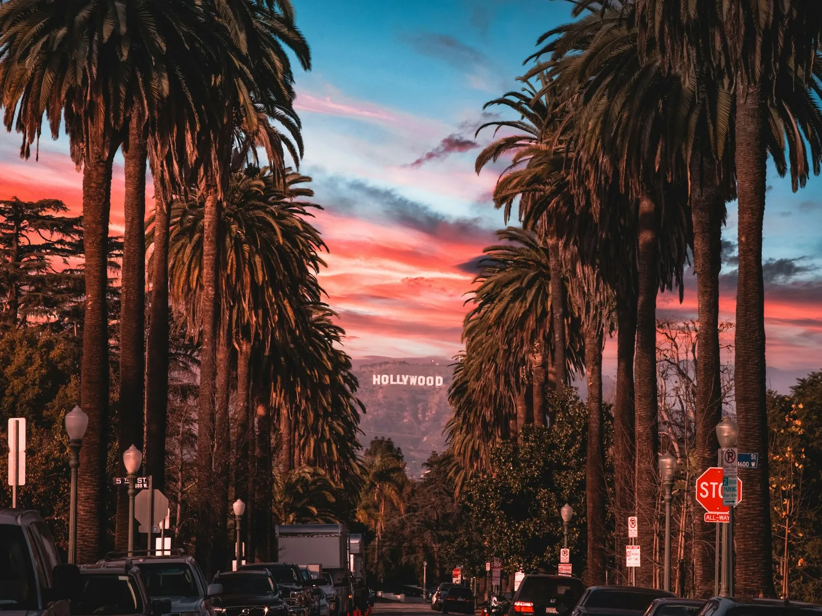 Rua de Hollywood ladeada por palmeiras altas ao entardecer, com o letreiro de Hollywood pequeno ao fundo