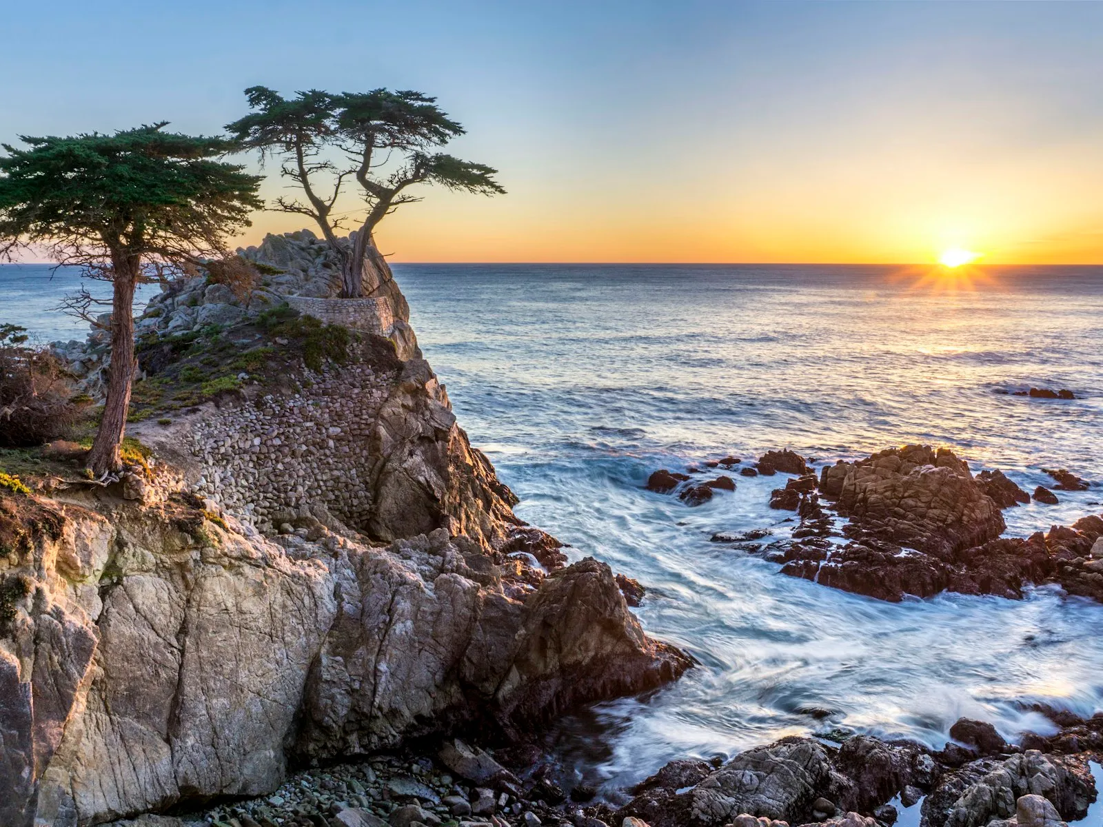O cipreste solitário (Lone Cypress) sobre a rocha da 17-Mile Drive ao pôr do sol, com ondas no Pacífico